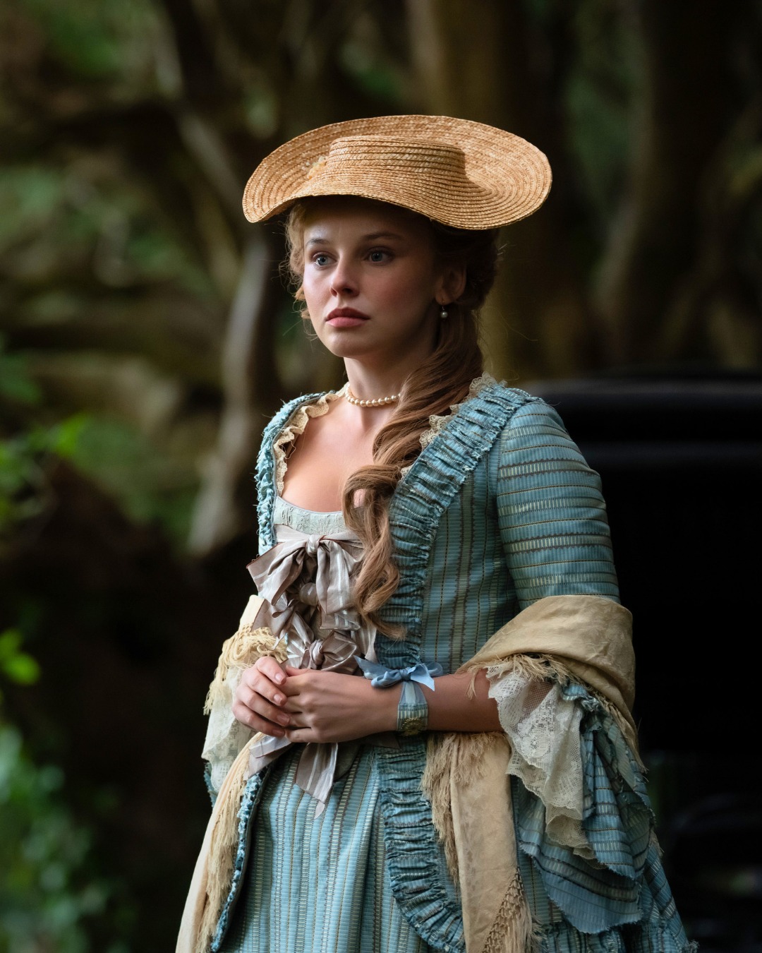 A young woman in an 18th-century blue gown and straw hat in a wooded estate.
