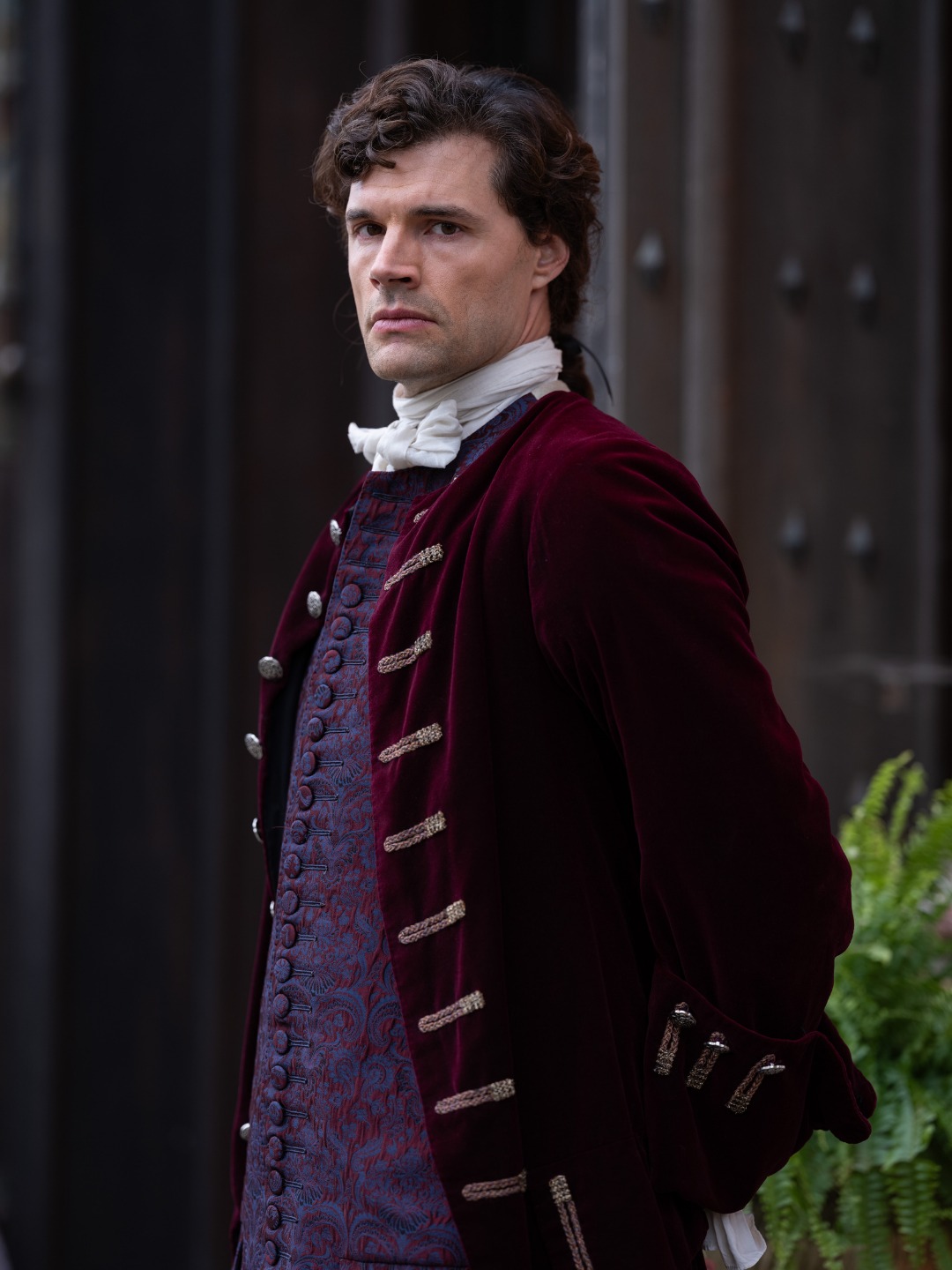 A nobleman in deep burgundy velvet standing before heavy estate doors.